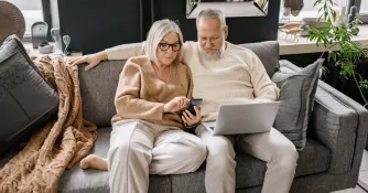 Äldre kvinna och man sitter i soffan med en laptop och smartphone.