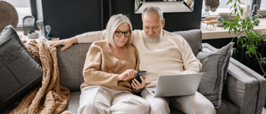 Äldre kvinna och man sitter i soffan med en laptop och smartphone.