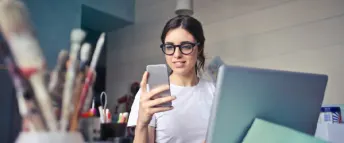 En kvinna sitter omgiven av penslar och pennor vid sin dator med en telefon i handen