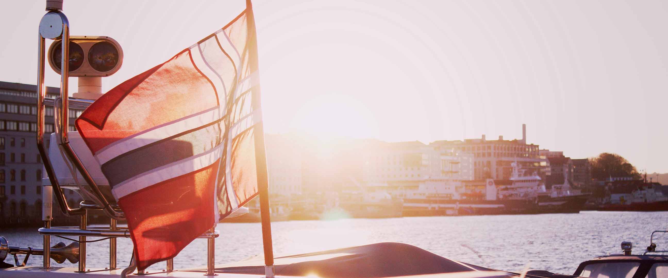Norsk flagga på båt i motljus, med stadsmiljö i bakgrunden