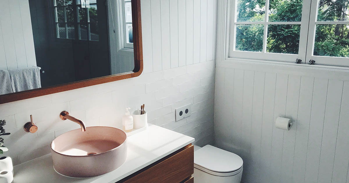 ett modernt badrum i vitt med kommod och spegel i trä samt toalett, dusch, fönster, handfat.
