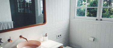 ett modernt badrum i vitt med kommod och spegel i trä samt toalett, dusch, fönster, handfat.