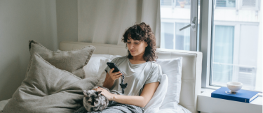 Kvinna sitter i sängen och kollar på mobilen tillsammans med en katt. 