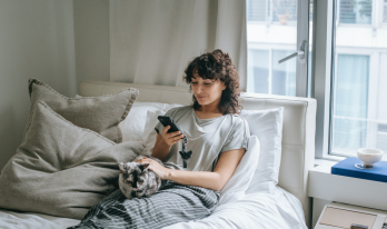 Kvinna sitter i sängen och kollar på mobilen tillsammans med en katt. 