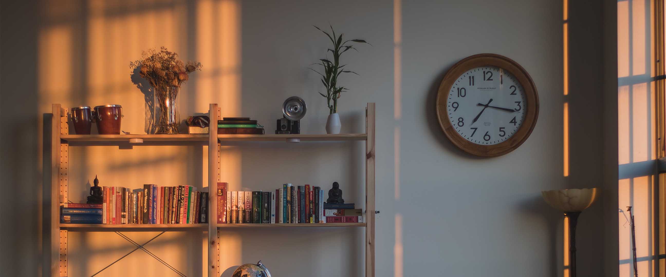 Klocka med träram på en vit vägg, bredvid en bokhylla full med böcker i olika färger