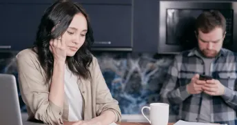 Kvinna sitter i ett kök och tittar på fakturor medan en man är på väg att ringa ett samtal i bakgrunden.