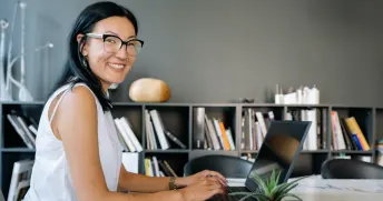 Ung kvinna med glasögon sitter vid en laptop och ler mot kameran.