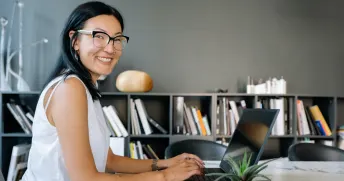 Ung kvinna med glasögon sitter vid en laptop och ler mot kameran.