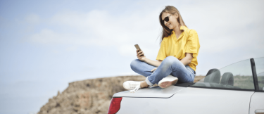 Kvinna sitter på en bil och kollar i mobilen