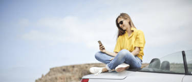 Kvinna sitter på en bil och kollar i mobilen