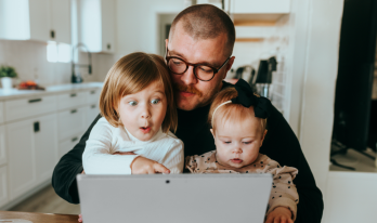 Pappa sitter med sina två småbarn framför en laptop i köket.