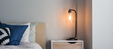 Bordslampa med glasskärm på nattduksbord intill bäddad säng med blå kudde.
