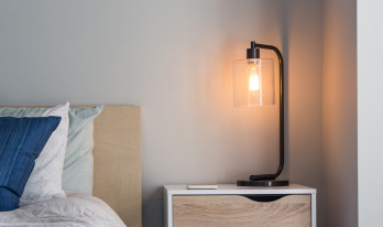 Bordslampa med glasskärm på nattduksbord intill bäddad säng med blå kudde.