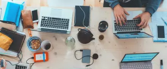 Flera datorer ligger på ett bord med te- och kaffekoppar, mobiler och andra saker runt omkring.