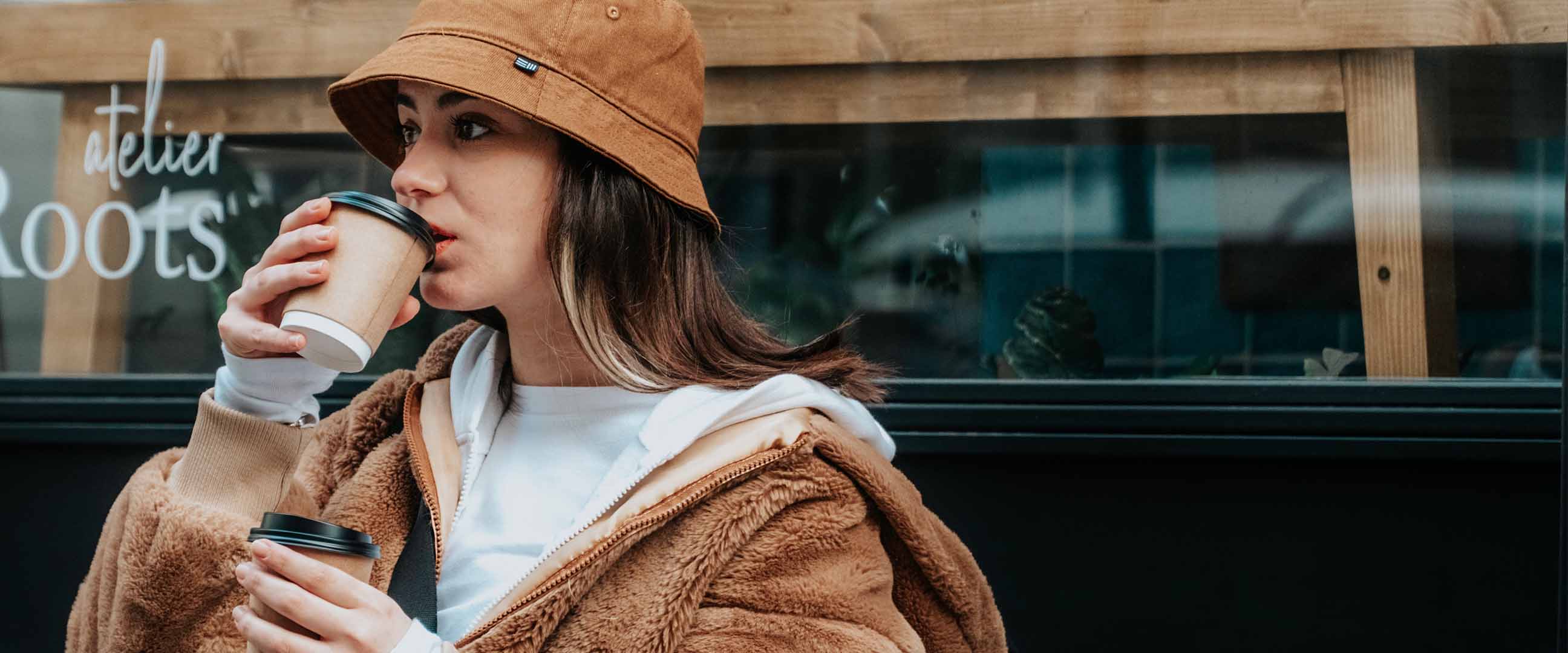 Kvinna i brun buckethat och hoodie dricker kaffe i take away-kopp.