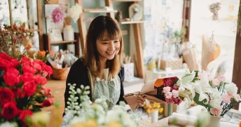 Florist som arbetar i sin egna blombutik, iförd ett ljusgrönt förkläde och håller i en bukett med vita och rosa blommor.