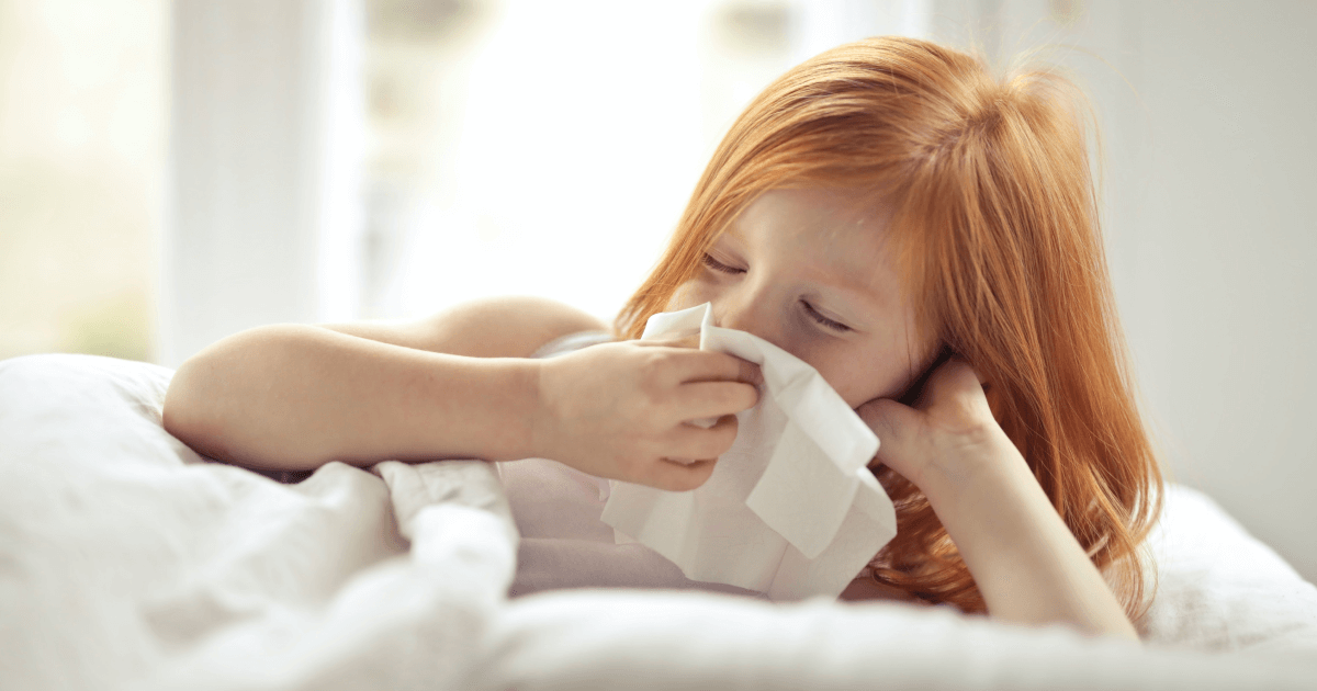 Förkyld rödhårig flicka ligger i sängen och torkar näsan med en näsduk. 