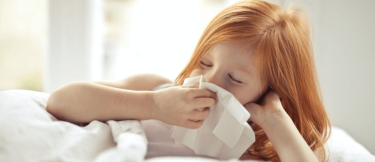 Förkyld rödhårig flicka ligger i sängen och torkar näsan med en näsduk. 