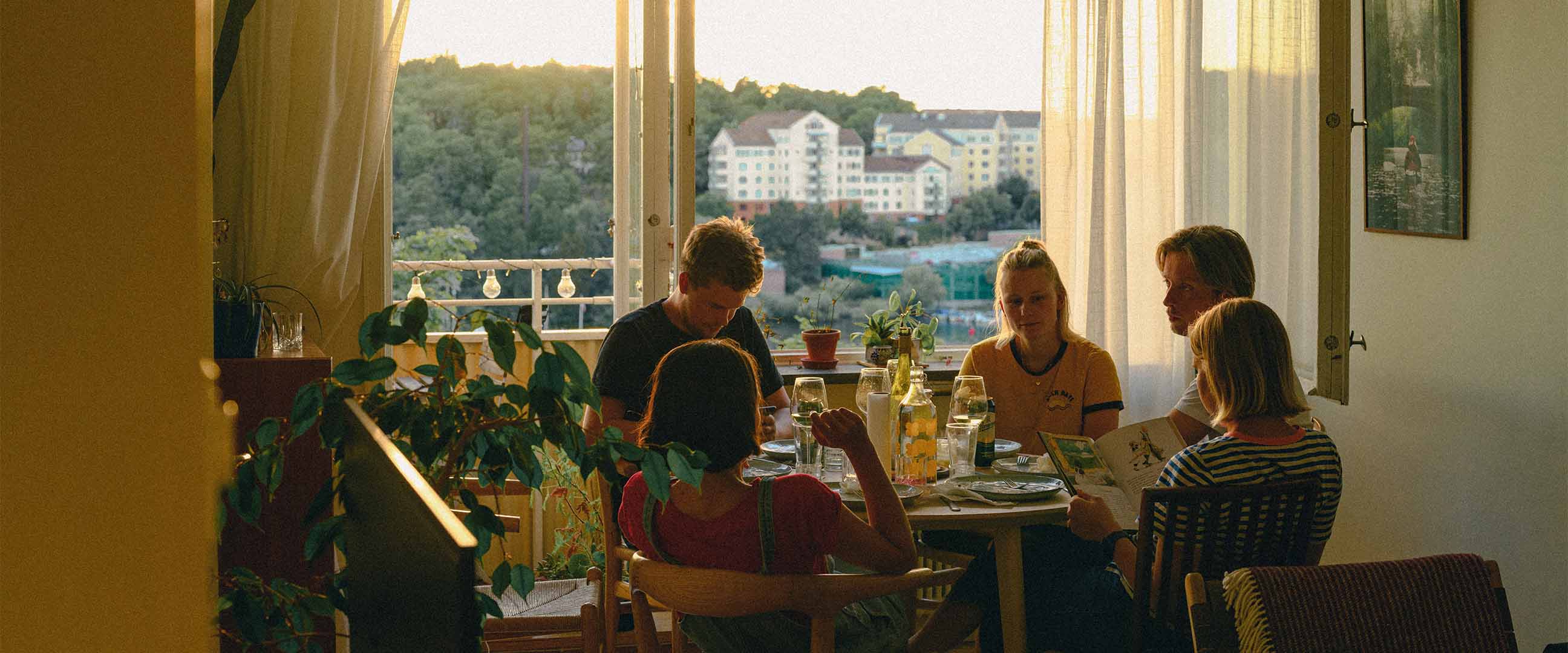 En grupp vänner som äter middag ihop hemma med balkongdörr öppen och utsikt över lägenhetshus