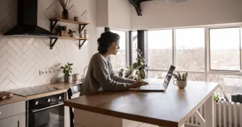 Kvinna sitter vid en köksö i ett vitt kök med stora fönster och skriver på en laptop. 