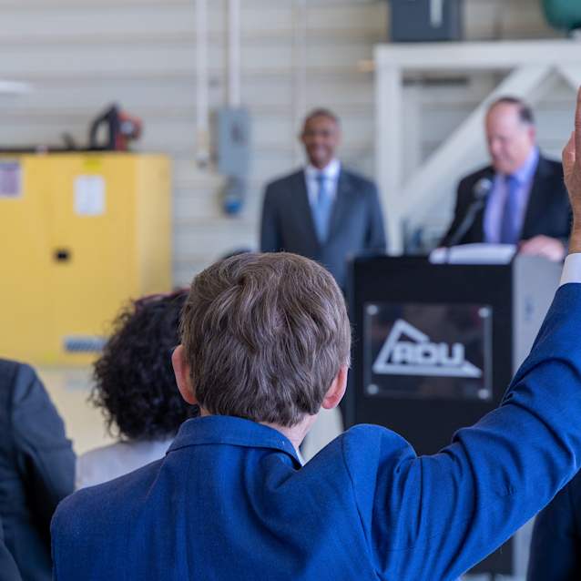 Person in blue suit raising hand during business presentation with speakers at podium and yellow cabinet in background.