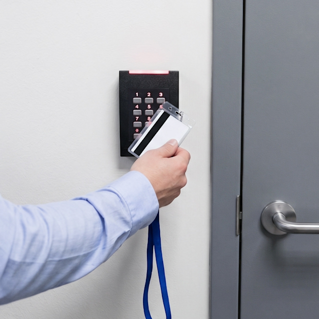 Person in light blue shirt holding white access card to black keypad reader mounted on white wall next to gray door.