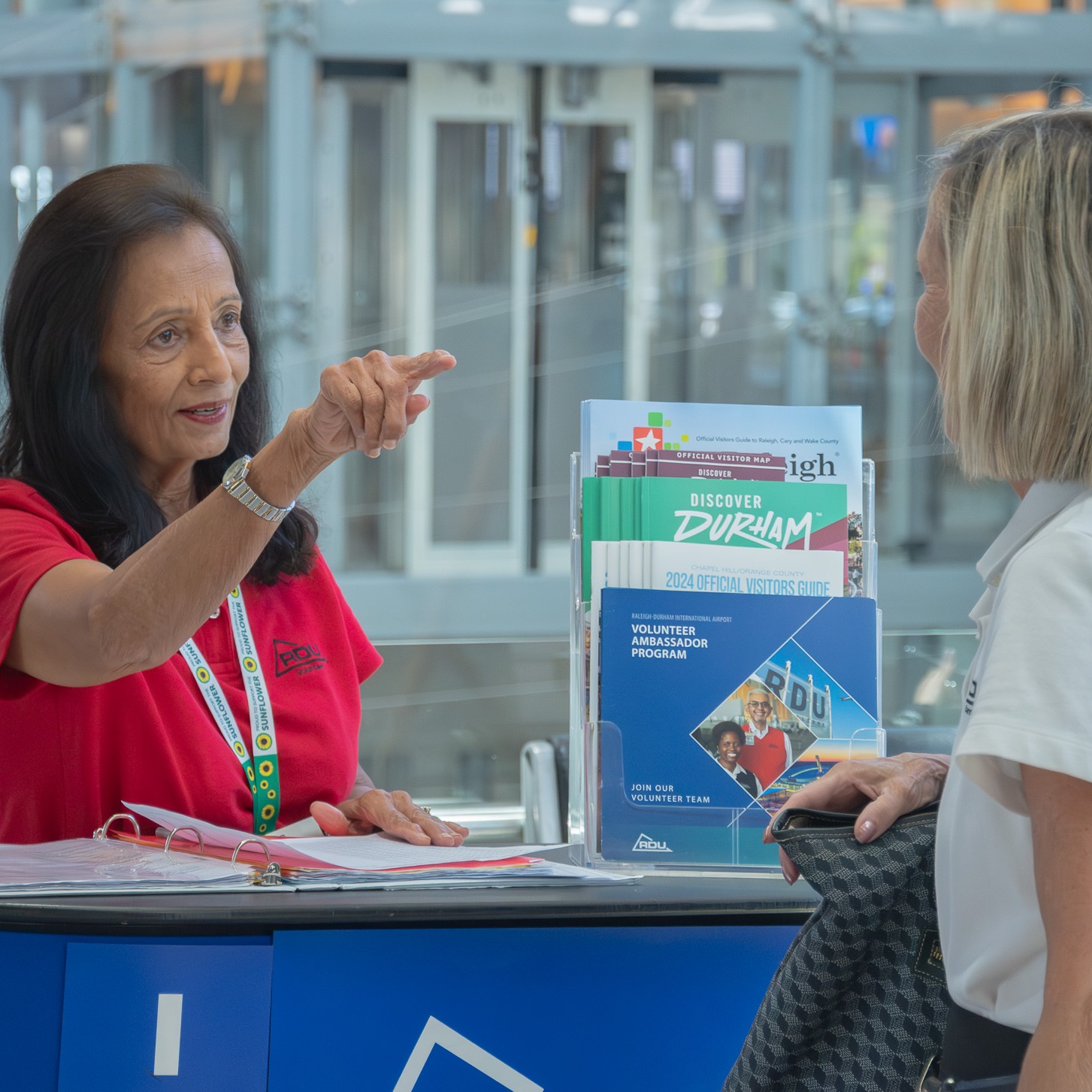 Volunteer in red shirt at information desk pointing and assisting a visitor with tourism brochures displayed.