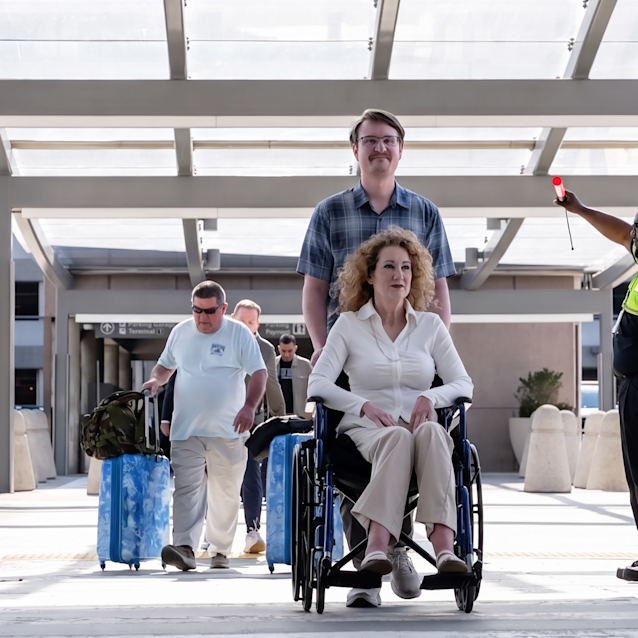 Man pushing woman in wheelchair at airport terminal with travelers and staff in background under modern canopy.