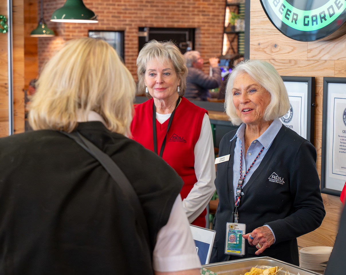 Two women in branded vests greet a visitor at a welcome desk in a modern space with brick walls and green pendant lights.