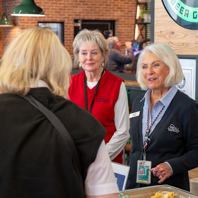 Two women in branded vests greet a visitor at a welcome desk in a modern space with brick walls and green pendant lights.