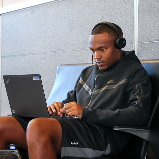 A guest wearing headphones and a hoodie works on a laptop while sitting near the airport gate.