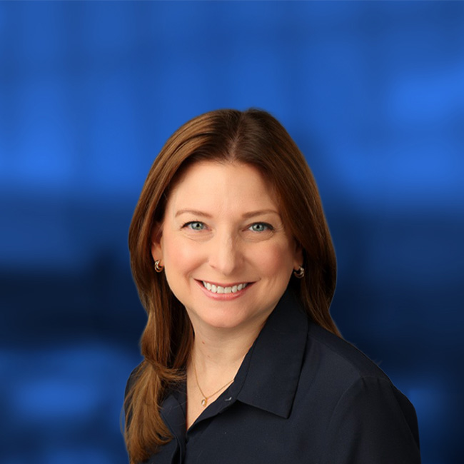 Professional headshot of a smiling woman with brown hair and green eyes wearing a black collared shirt against a blue background.