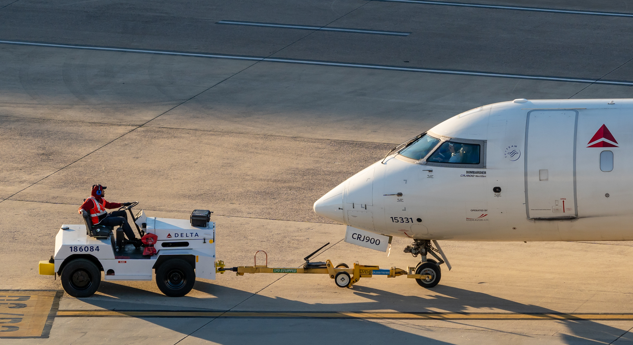 Ground crew member in safety vest operates white Delta tug vehicle connected to nose of Delta CRJ900 aircraft on tarmac.