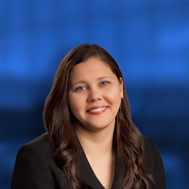 Professional headshot of a smiling woman with long brown hair wearing a black blazer against a blue background.