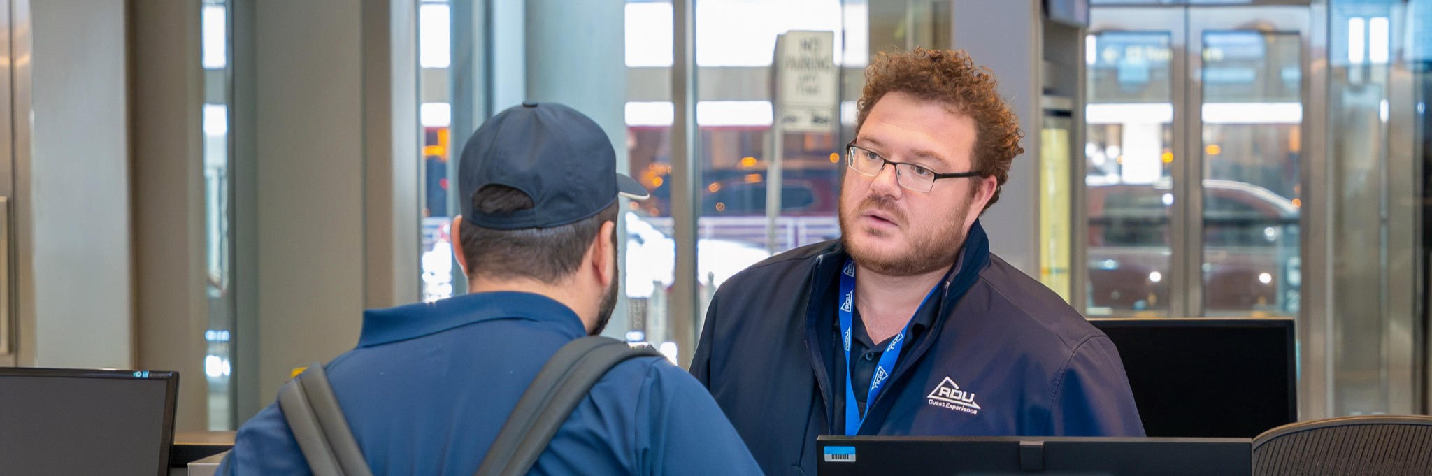 Staff member in navy jacket and glasses assists customer with backpack at service counter in modern facility.