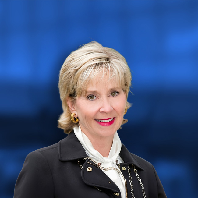 Professional headshot of a blonde woman in a black jacket with gold accents and white blouse against a blue background.
