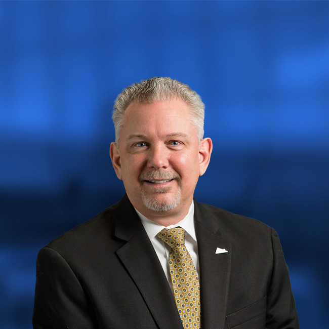 Professional headshot of a smiling man in a dark suit with yellow patterned tie and white pocket square against a blue background.