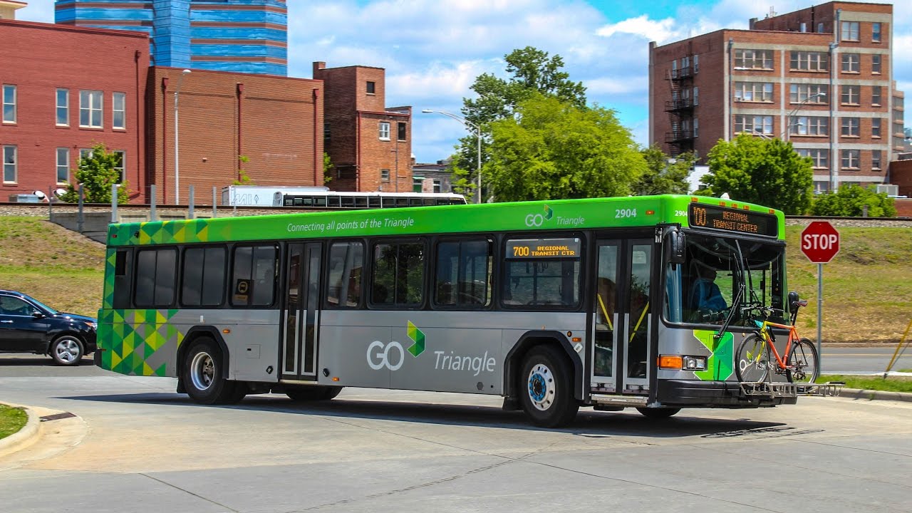 GO Triangle public transit bus in green and gray livery with bike rack, traveling on city street with buildings behind.