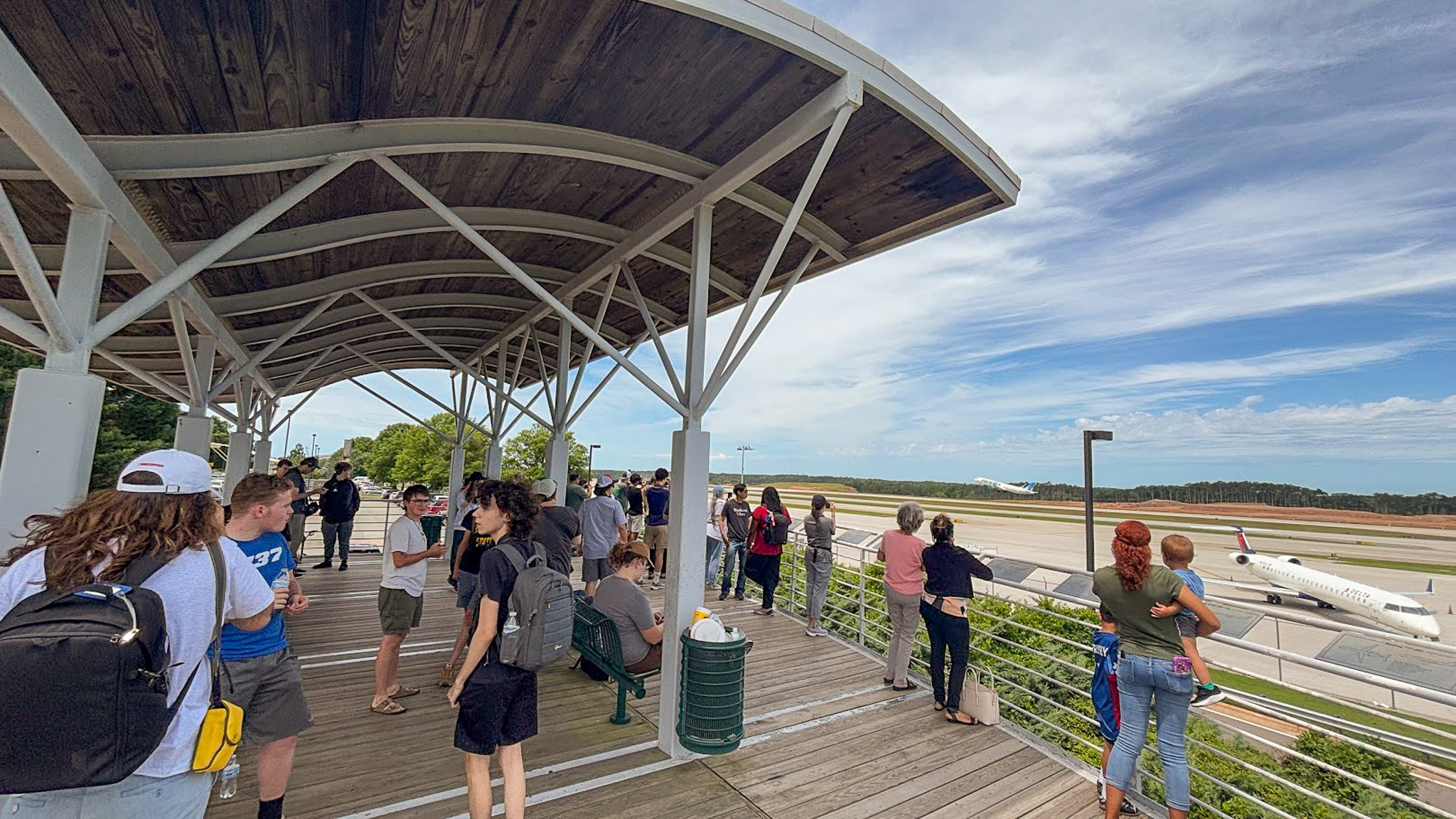 People gathered under a covered observation deck viewing an airport runway with an airplane under blue skies.