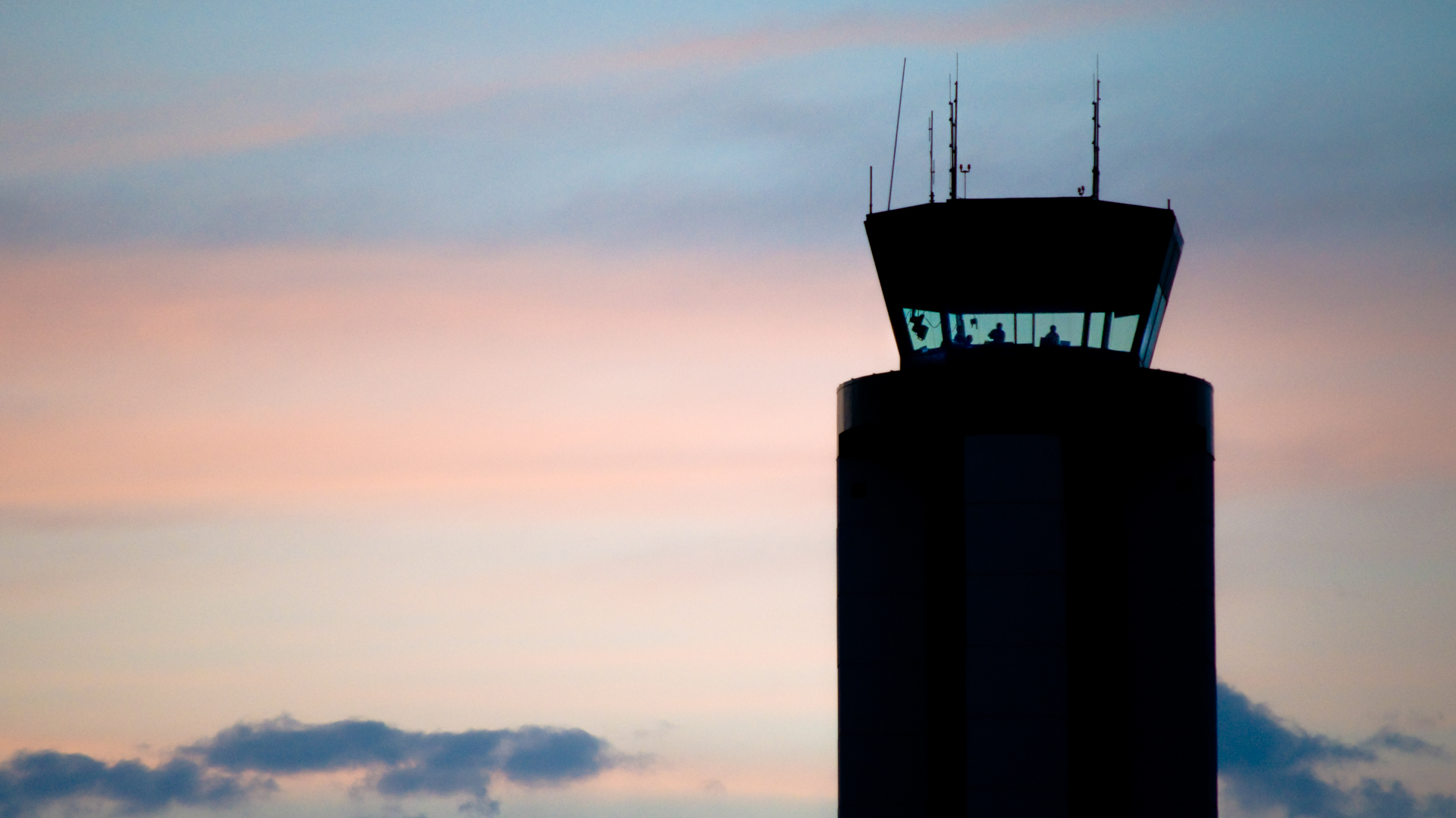 Silhouette of airport control tower with illuminated windows showing air traffic controllers at dusk with pink and blue sky.