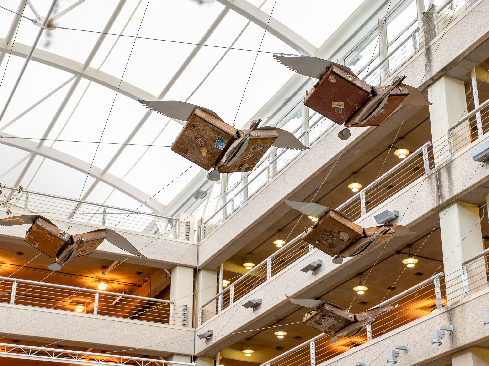 Vintage suitcases with wings suspended from glass ceiling in modern multi-story atrium with balconies and warm lighting.