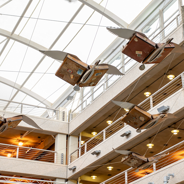 Vintage suitcases with wings suspended from glass ceiling in modern multi-story atrium with balconies and warm lighting.