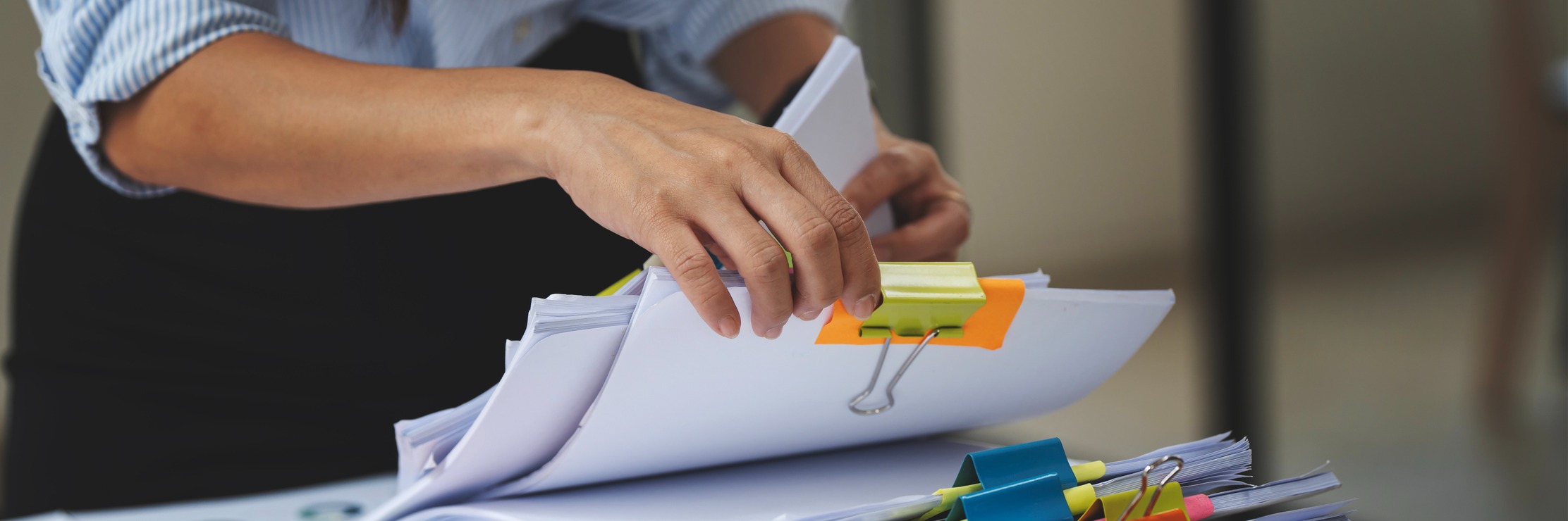 Person in blue striped shirt organizing stacks of white papers with colorful binder clips and sticky notes on desk.
