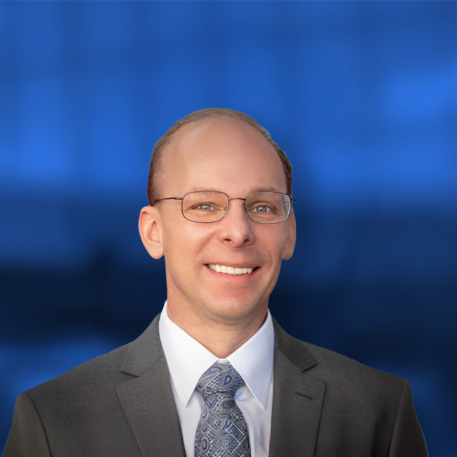 Professional headshot of a smiling man wearing glasses, a dark suit, white shirt, and patterned blue tie against a blue background.