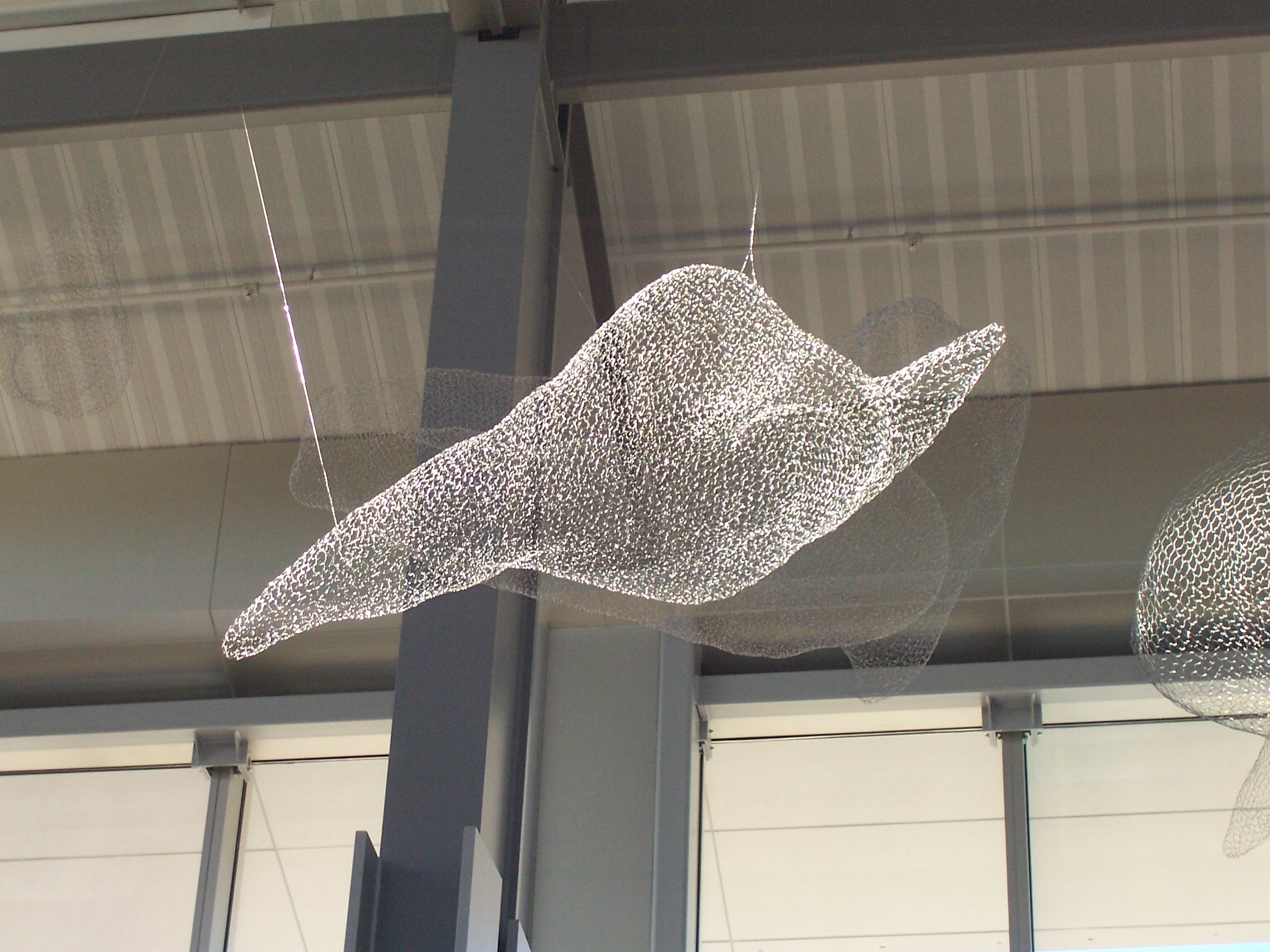 Wire mesh sculpture of a cloud suspended from an industrial ceiling with metal beams and white walls visible.