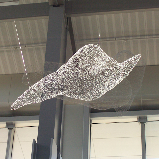 Wire mesh sculpture of a cloud suspended from an industrial ceiling with metal beams and white walls visible.