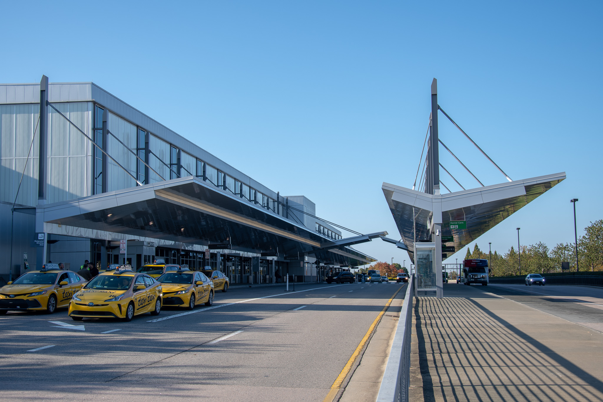 Modern airport terminal with yellow taxis lined up outside under a clear blue sky and distinctive angular architecture.