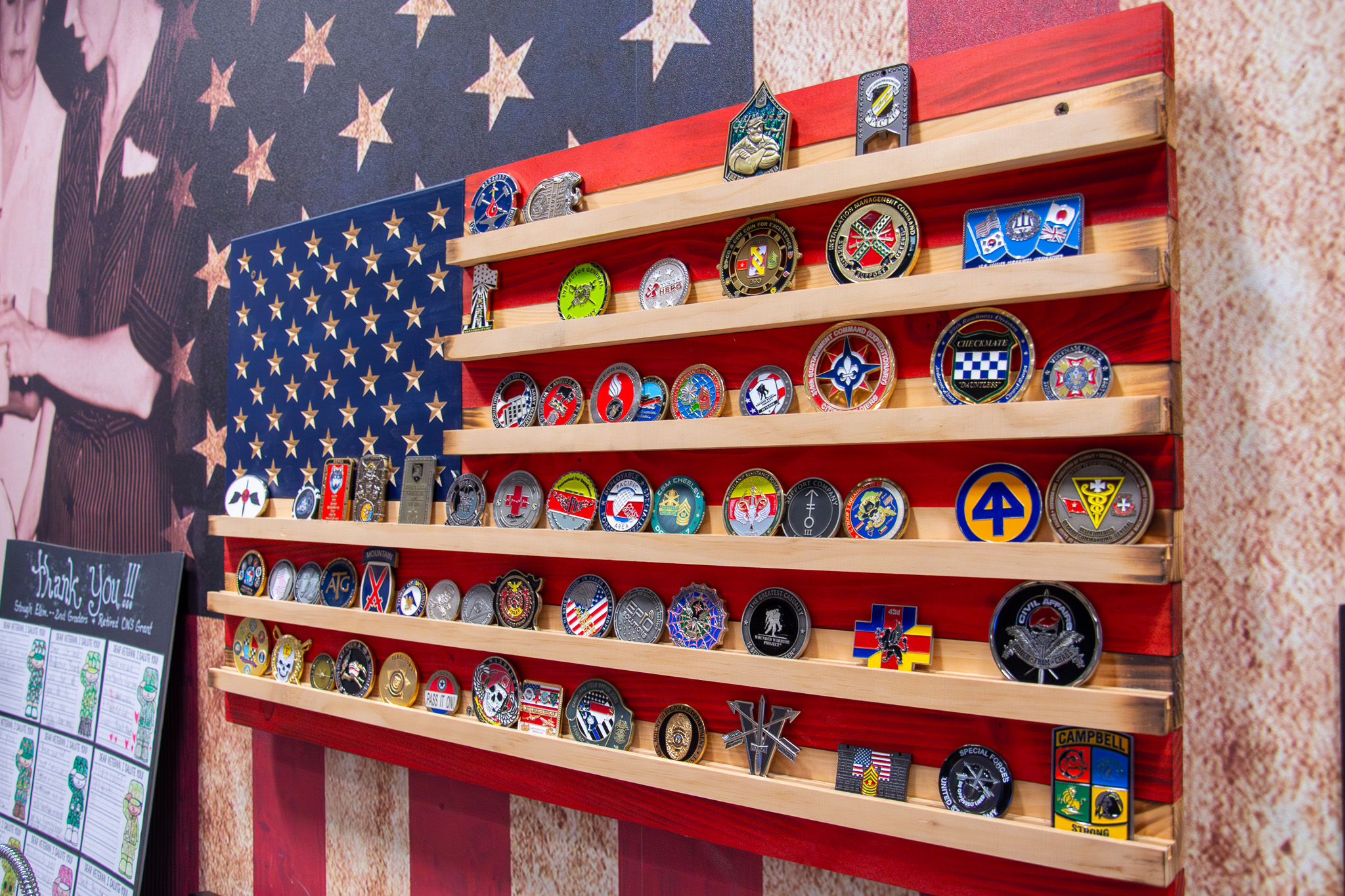 Military challenge coins displayed on wooden shelves arranged in the pattern of an American flag with red and white stripes.