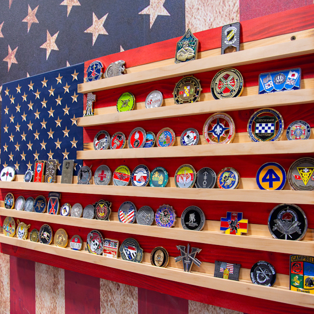 Military challenge coins displayed on wooden shelves arranged in the pattern of an American flag with red and white stripes.
