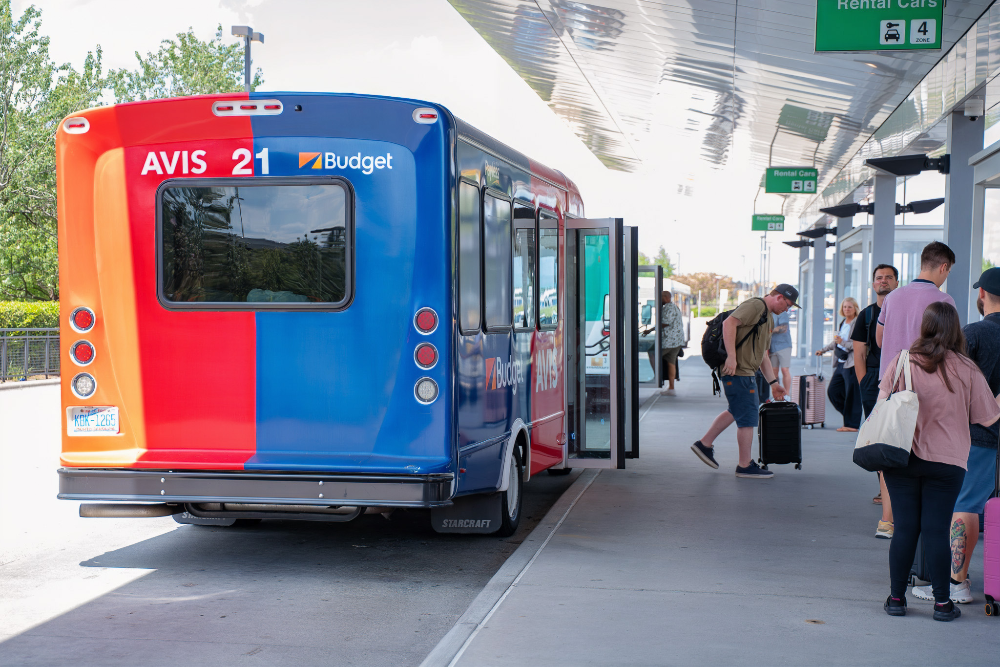 Red and blue Avis Budget shuttle bus number 21 loading passengers with luggage at airport rental car terminal.